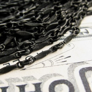 Close-up view of a pile of black metal chains. The chains have a textured, ridged surface and are linked together. The chains appear to be for jewelry making, with small, uniform links. The overall color is a matte black.