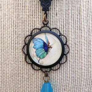 May include: A necklace with a blue butterfly charm and a teardrop-shaped blue glass pendant. The butterfly charm is set in a round, black frame with a copper-colored filigree border.