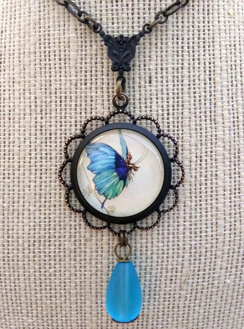 May include: A necklace with a blue butterfly charm and a teardrop-shaped blue glass pendant. The butterfly charm is set in a round, black frame with a copper-colored filigree border.