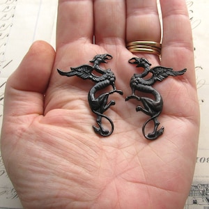 Op de afbeelding: Twee zwarte metalen griffin-beeldjes met uitgespreide vleugels. De griffioenen staan ​​tegenover elkaar en staan ​​op een hand.