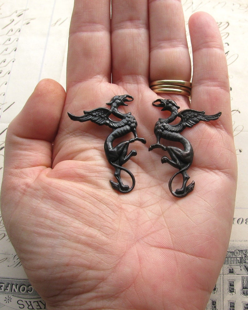 Op de afbeelding: Twee zwarte metalen griffin-beeldjes met uitgespreide vleugels. De griffioenen staan ​​tegenover elkaar en staan ​​op een hand.
