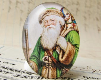 May include: An oval-shaped Christmas ornament featuring a vintage illustration of Santa Claus. Santa is depicted with a long white beard, wearing a green coat with brown fur trim, and carrying a sack over his shoulder. He holds a wooden staff.