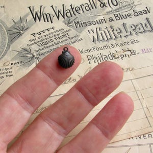 May include: A small, dark gray, metal seashell charm. The charm is being held by a finger.