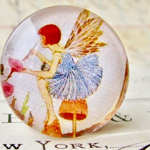 May include: A glass cabochon with a vintage illustration of a fairy with brown wings and a blue dress standing on a mushroom. The fairy is holding a flower in her hand.