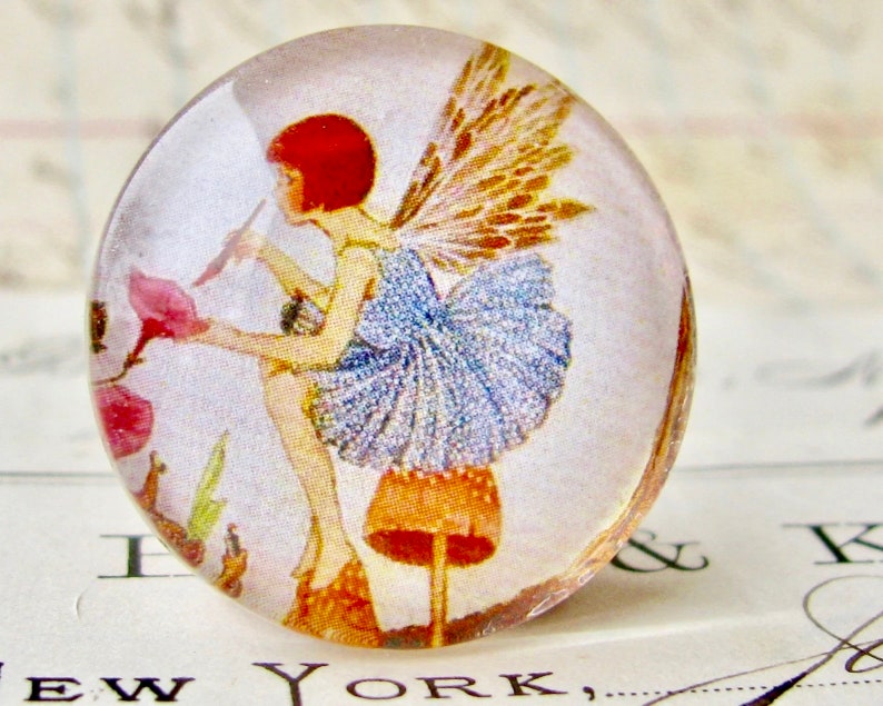 May include: A glass cabochon with a vintage illustration of a fairy with brown wings and a blue dress standing on a mushroom. The fairy is holding a flower in her hand.