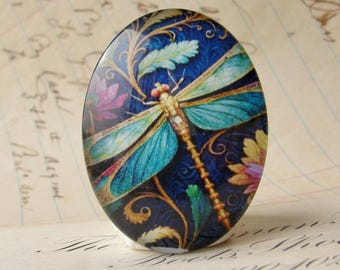 Op de afbeelding: Ovale decoratieve glazen cabochon met een libellenontwerp. De libel heeft turquoise vleugels en een gouden lichaam, tegen een donkerblauwe achtergrond met bloemaccenten in roze, geel en groen.