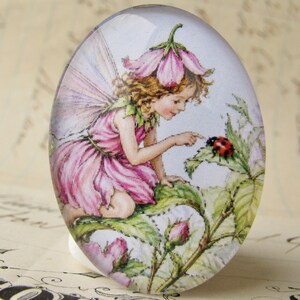 May include: An oval glass cabochon featuring a detailed illustration of a fairy with light brown hair, wearing a pink dress and a flower hat, sitting on green leaves and reaching out to touch a small red and black ladybug on a leaf.