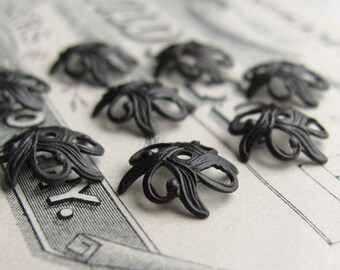 May include: A group of eight black metal decorative embellishments with a floral design. The embellishments are small and intricate, with a delicate, filigree-like pattern.
