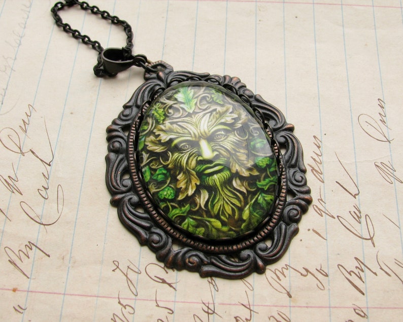 Puede incluir: Un collar con un colgante de bronce con un camafeo de vidrio verde y marr&oacute;n que presenta un rostro de hombre verde rodeado de hojas.