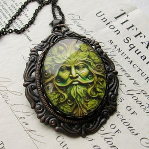 Op de afbeelding: Een bronzen ketting met een hanger met een groene man ontwerp. De hanger heeft een ronde glazen cameo met een gedetailleerd beeld van een groene man met gezicht, omringd door bladeren. De ketting heeft een zwarte ketting.