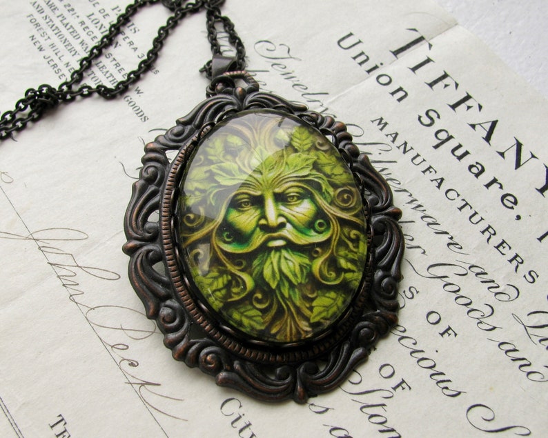 Op de afbeelding: Een bronzen ketting met een hanger met een groene man ontwerp. De hanger heeft een ronde glazen cameo met een gedetailleerd beeld van een groene man met gezicht, omringd door bladeren. De ketting heeft een zwarte ketting.