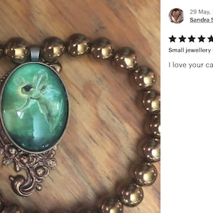Op de afbeelding: Een koperen ketting met een ovale hanger in groen en goud met een afbeelding van een fee. De ketting is gemaakt met ronde, metalen kralen. De hanger is 40 millimeter lang. "Kleine sieradenlengte: 40 millimeter Ik hou van je cabochons! Dank je!"