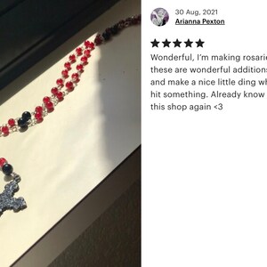 May include: A rosary with red and black beads, a silver-tone cross pendant, and a small oval pendant with an image. The rosary is displayed on a light-colored surface.