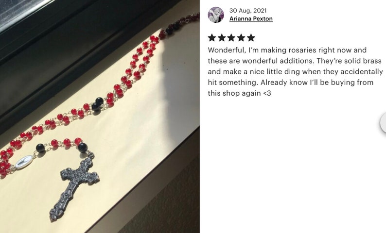 May include: A rosary with red and black beads, a silver-tone cross pendant, and a small oval pendant with an image. The rosary is displayed on a light-colored surface.