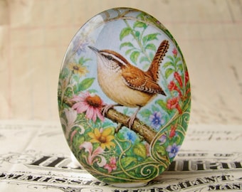 May include: Oval-shaped decorative object featuring a detailed illustration of a bird perched on a branch. The bird has brown and tan feathers, surrounded by colorful flowers and foliage against a light blue background. The object has a glossy finish.