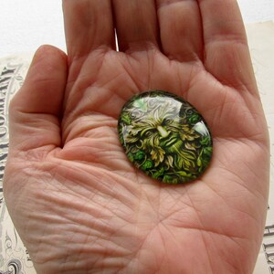 Puede incluir: Un cabuj&oacute;n de vidrio verde con una imagen detallada del Hombre Verde, una figura m&iacute;tica a menudo asociada con la naturaleza y la fertilidad.