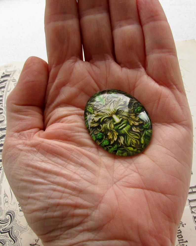 Puede incluir: Un cabuj&oacute;n de vidrio verde con una imagen detallada del Hombre Verde, una figura m&iacute;tica a menudo asociada con la naturaleza y la fertilidad.