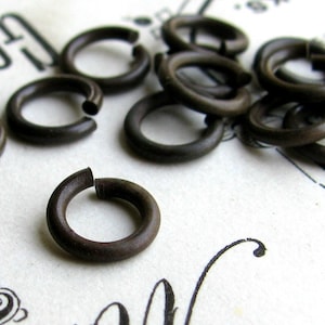 K&ouml;nnte beinhalten: Eine Sammlung kleiner, dunkelbrauner Metall-Sprungringe. Die Ringe sind offen, mit einer kleinen L&uuml;cke, an der sie mit anderen Objekten verbunden werden k&ouml;nnen. Sie liegen auf einer wei&szlig;en Oberfl&auml;che mit verblasstem schwarzem Text und dekorativen Elementen.