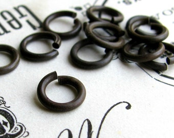 May include: A collection of small, dark brown metal jump rings. The rings are open, with a small gap where they can be connected to other objects. The rings are scattered on a white surface with faded black text and decorative elements.