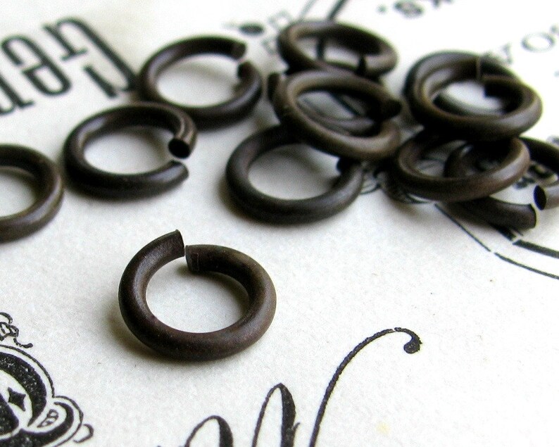 K&ouml;nnte beinhalten: Eine Sammlung kleiner, dunkelbrauner Metall-Sprungringe. Die Ringe sind offen, mit einer kleinen L&uuml;cke, an der sie mit anderen Objekten verbunden werden k&ouml;nnen. Sie liegen auf einer wei&szlig;en Oberfl&auml;che mit verblasstem schwarzem Text und dekorativen Elementen.