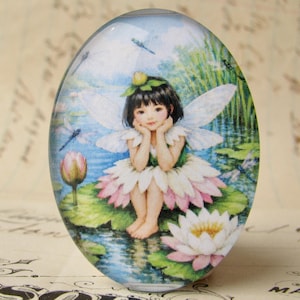 May include: Oval-shaped glass paperweight featuring a fairy illustration. The fairy has black hair, white wings, and a flower dress, sitting on a lily pad. The background shows a pond with dragonflies and water lilies.
