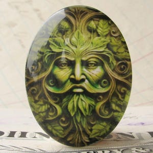 Op de afbeelding: Een ovale glazen cabochon met een gedetailleerd beeld van de Green Man, een mythische figuur met een gezicht omringd door bladeren en wijnstokken, in tinten groen en bruin.