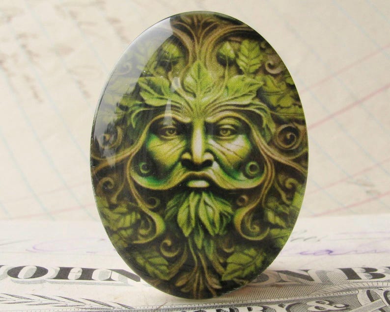 Op de afbeelding: Een ovale glazen cabochon met een gedetailleerd beeld van de Green Man, een mythische figuur met een gezicht omringd door bladeren en wijnstokken, in tinten groen en bruin.