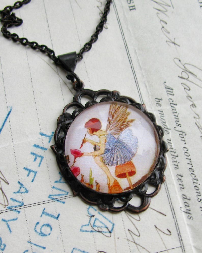 May include: A copper-toned pendant necklace with a glass cameo featuring a fairy with blue wings sitting on a mushroom. The fairy is holding a flower and is surrounded by other mushrooms and greenery. The cameo is set in a filigree frame.