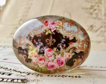 May include: Oval-shaped glass cabochon with a floral design. The design features pink roses, a fleur-de-lis, and ornate scrollwork against a black and light blue background. The cabochon has a slightly domed surface.