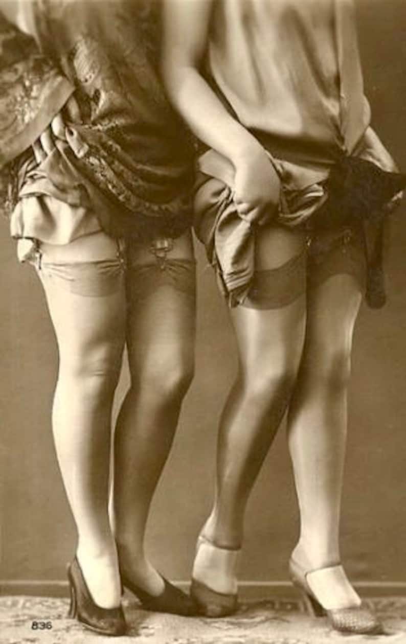 May include: Two women wearing stockings and shoes. The women are standing close together and their legs are crossed. The photo is in black and white.