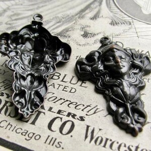 May include: Two black metal pendants with intricate designs. One pendant features a woman's face with flowing hair and the other pendant features a floral design.