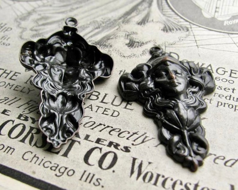 May include: Two black metal pendants with intricate designs. One pendant features a woman's face with flowing hair and the other pendant features a floral design.