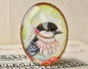 May include: Oval-shaped glass cabochon featuring a watercolor illustration of a bird with a black and white head, a pink flower, and a pink and white collar. The bird is set against a green and yellow background, framed by a brown border.