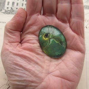 Op de afbeelding: Een groene glazen cabochon met een geel en groen fee&euml;nontwerp. De cabochon is ovaalvormig en heeft een glad, glanzend oppervlak.