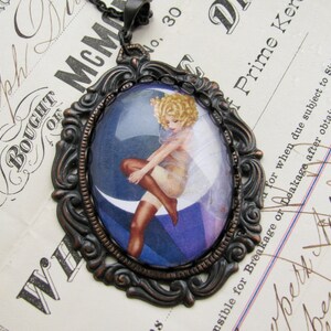 May include: A vintage-style pendant necklace with a cameo featuring a woman in a white dress and stockings, sitting on a crescent moon. The cameo is set in an ornate bronze-coloured frame.