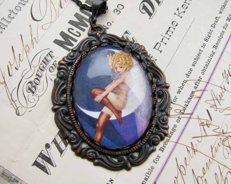 May include: A vintage-style pendant necklace with a cameo featuring a woman in a white dress and stockings, sitting on a crescent moon. The cameo is set in an ornate bronze-coloured frame.