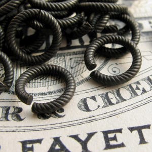 May include: A pile of black metal jump rings, each with a twisted rope design. The rings are scattered on a white surface with black text.