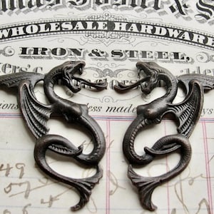 May include: Two antique bronze dragon-shaped metal embellishments with wings and tails. The dragons are facing each other with their heads turned to the side. The embellishments are on a white background with black text.