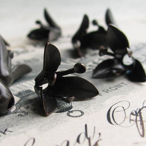May include: A set of six black metal flower charms with a vintage look. The charms are in the shape of a flower with five petals and a small ball in the center. The charms are arranged on a white surface with the word "Front" printed on it.