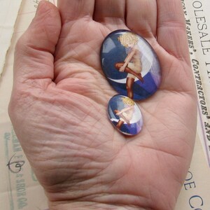 May include: Two glass cabochons with a blue background and a woman in a white dress standing on a crescent moon. The larger cabochon is about 5 cm in diameter and the smaller cabochon is about 2.5 cm in diameter.