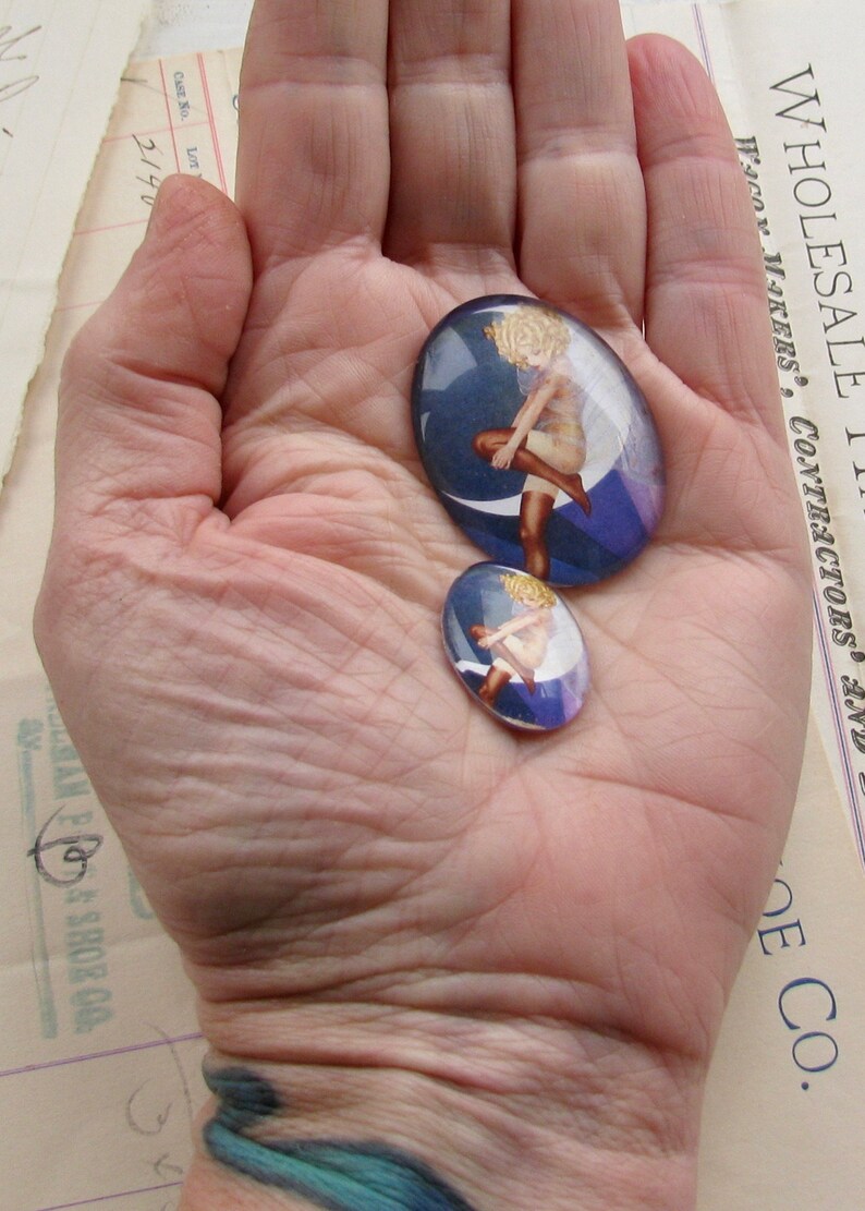 May include: Two glass cabochons with a blue background and a woman in a white dress standing on a crescent moon. The larger cabochon is about 5 cm in diameter and the smaller cabochon is about 2.5 cm in diameter.
