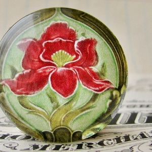 May include: A round glass paperweight with a floral design. The paperweight features a red flower with green leaves and a brown border.