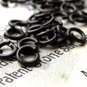 May include: A close-up of a pile of black metal jump rings. The rings are small and round, and they are scattered on a white surface.