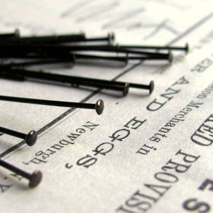 May include: A close-up of a pile of black straight pins with round heads. The pins are scattered on a piece of paper with text that reads "Smoked Provisions", "New York", and "Edge & Co."