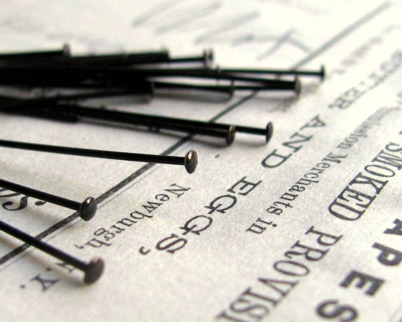 May include: A close-up of a pile of black straight pins with round heads. The pins are scattered on a piece of paper with text that reads "Smoked Provisions", "New York", and "Edge & Co."