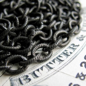 May include: A close-up of a black metal chain with a textured surface. The chain is lying on a piece of paper with the text "310MMR & CO" printed on it.