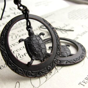 May include: A pair of black oxidized silver earrings with a detailed floral design. Each earring features a small turtle charm hanging from the center.