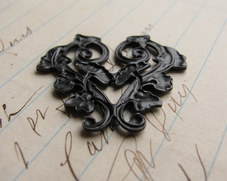 May include: Black metal heart-shaped decorative embellishment with a floral design. The embellishment is on a sheet of lined paper.