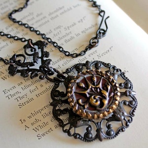 May include: A black metal necklace with a large, ornate pendant. The pendant features a round, red and gold button with a floral design. The chain is made of small, linked rings.