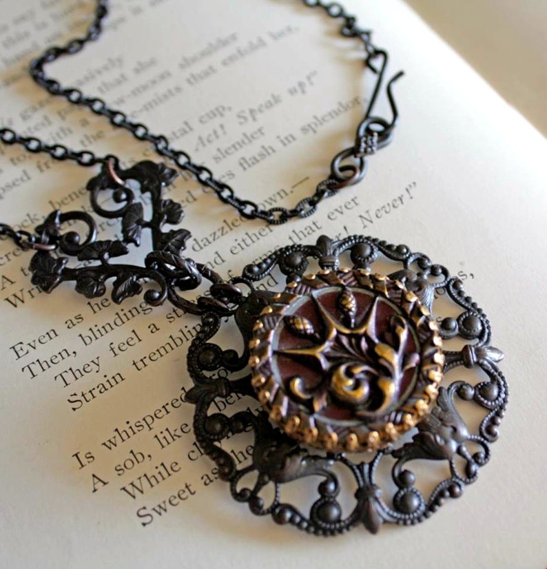 May include: A black metal necklace with a large, ornate pendant. The pendant features a round, red and gold button with a floral design. The chain is made of small, linked rings.